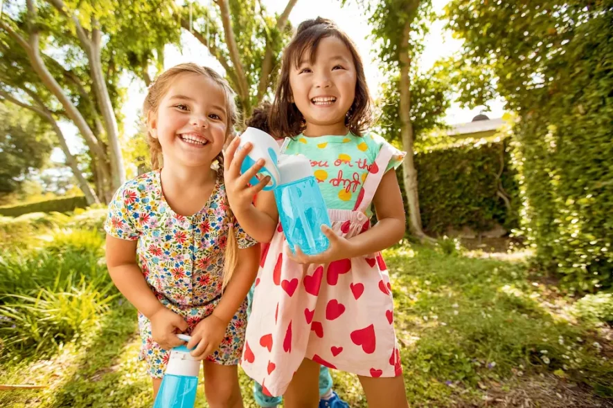 Petites filles heureuses avec gourdes écologiques, rappelant l'importance de boire régulièrement pour les enfants en été.